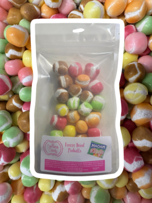 A package of freeze-dried pinballs candy displayed in front of a blurred background of multicolored candies. The front package is clear, showing a variety of freeze-dried candies in hues of brown, pink, yellow, and green, with a label